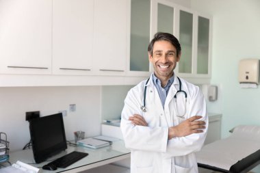 Kendine güvenen bir pratisyen. Gülümseyen mutlu erkek doktor portresi modern sağlık merkezinin iyi donanımlı muayene odasında poz veriyor.
