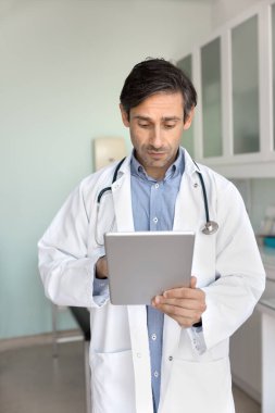 Doktor servisinde modern teknoloji. Profesyonel orta yaşlı erkek hekim hastane ofisinde dokunmatik ekranlı cihaz üzerinde çalışıyor. Tıbbi kaynakları internette tarıyor ve elektronik hasta kayıtlarını yönetiyor.