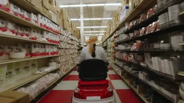 Mujer con una discapacidad en la silla de ruedas scooter de movilidad eléctrica elige los productos en la fila de compras en el supermercado, vista desde atrás. 4k . — Vídeos de Stock