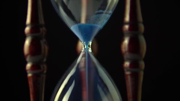 Rotating hourglass clock time concept. Video opening sand time ending at the end. Old vintage wood frame blue sand clock slowly spinning on dark background. Close up.