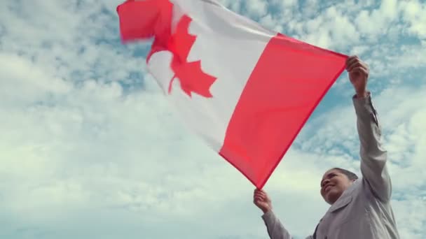 風でカナダの旗を掲げるアフリカ系アメリカ人女性の横からの肖像画で、太陽と青い曇りの空を見ている。幸せなカナダ市民。愛国者の概念. — ストック動画
