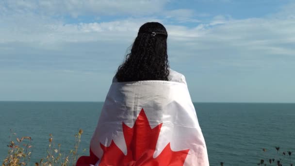 Afro-Amerikaanse Canadese volwassen vrouw met vlag als cape, ruitje of deken op haar schouders en zittend of kijkend naar het Ontario meer vanaf de klif, in de zomer zonnige, gouden uur avond. — Stockvideo