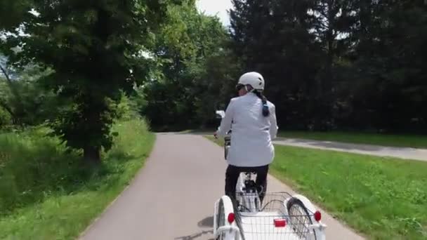 Güneşli yaz gününde parkta elektrikli bisiklete binen köylü kadın. Sırtından vurulmuş. Doğal ışıklandırma. Arkadan güç bataryasının görüntüsü. Yaz tatili yaşam tarzı. — Stok video