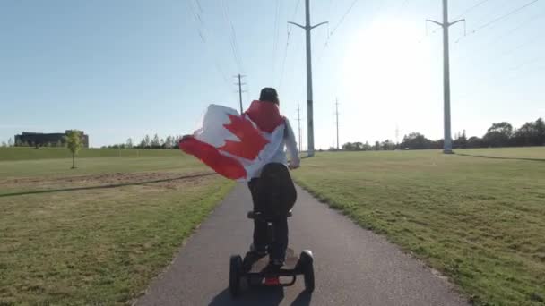 Femmes conduisant un scooter électrique dans le parc de la ville tout en tenant le drapeau canadien au vent. Citoyen canadien heureux conduisant un transport écologique et respectueux de l'environnement. Mode de vie adulte actif. — Video