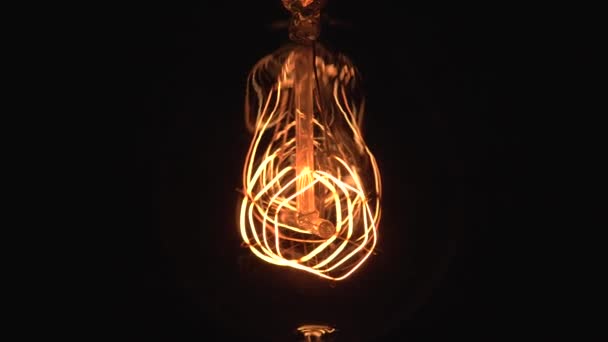 La lampada a lampadina incandescente Edison con un filamento di tungsteno illuminano e si muovono oscillando sul filo. Luce gialla su sfondo nero, vista da vicino. 4k . — Video Stock