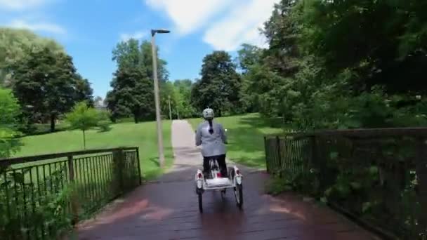 Donna in casco cavalcando bicicletta elettrica nel parco in soleggiata giornata estiva. Colpo alla schiena. Illuminazione naturale. La vista della batteria di alimentazione da dietro. Stile di vita di svago estivo. — Video Stock