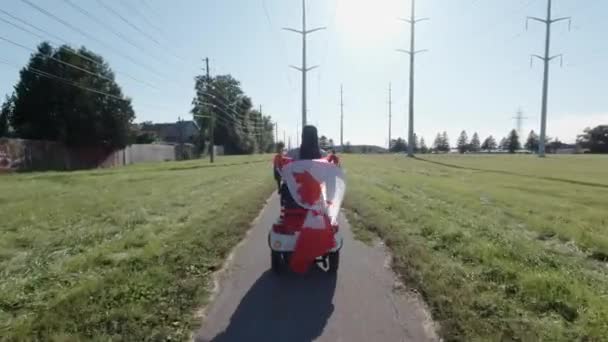 A nők elektromos robogón lovagolnak a városi parkban, miközben kanadai zászlót tartanak a szélben. Boldog kanadai állampolgár, aki környezettudatos energiaszállítást vezet. Aktív felnőtt életmód. — Stock videók