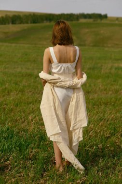 Çimlerin üzerinde güzel piknik. Yeşil tarlanın ortasında güzel, kızıl saçlı bir kadın iyi vakit geçiriyor. Yaz manzarası, güzel hava. Güneşli ve bulutlu rüzgarlı bir gün. Beyaz pamuk elbise eko tarzı