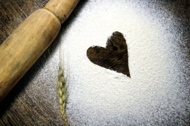 Buğday ununun arka planında toplanan taze buğday taneleri. Ahşap arka planda pişirme konsepti, kalple serpiştirilmiş un. Ahşap zemin üzerinde ahşap bir raptiye ile dağınık un içinde kalp çizilmiş