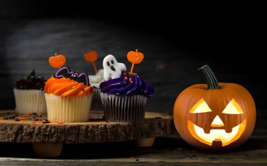 Mutlu Cadılar Bayramı. Balkabağı Jack-o-fener. Kek tatlısı. Renkli krema, buzlu hayalet ve sopayla süslenmiş. Cadılar Bayramı partisi. Ahşap arka planda kapkekler. 