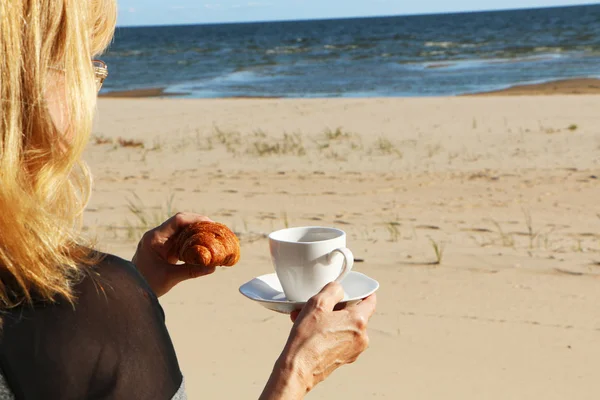 Meisje met een croissant en een kop koffie aan zee — Stockfoto