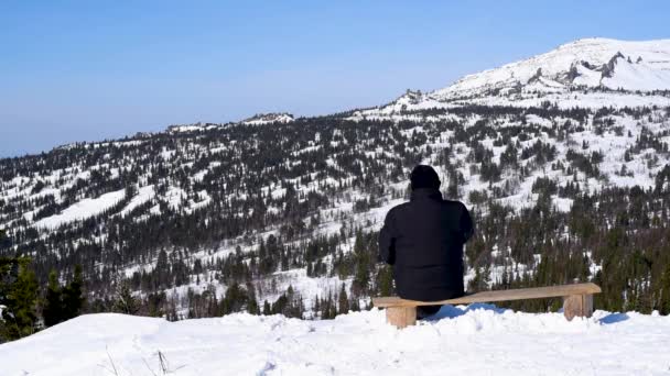 美しい雪景色を眺めながらスキー場のベンチに座った男が 晴天の冬の雪山を背景にたばこを吸う — ストック動画