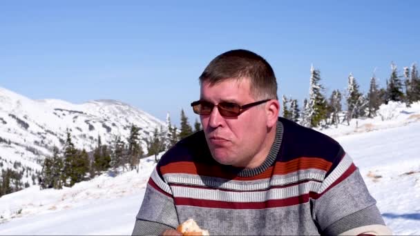 スキーリゾートの男が食べる 雪山と青空を背景に暖かい天候の冬はスキーリゾートでハンバーガーを食べる男 晴れの天気クローズアップで冬のスキーリゾートでピクニック — ストック動画