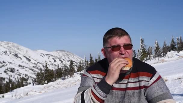 雪山と青空を背景に暖かい天候の中 冬はスキー場でハンバーガーを食べる男 — ストック動画