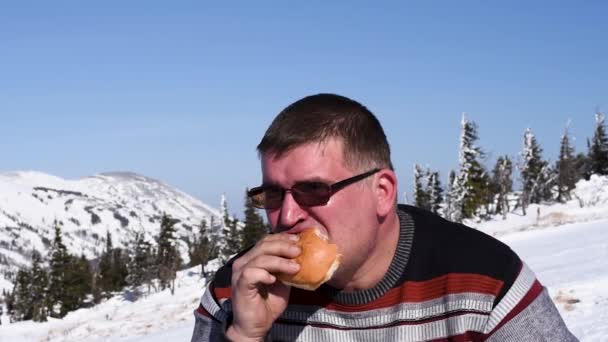 雪山と青空を背景に暖かい天候の中 冬はスキー場でハンバーガーを食べる男 — ストック動画