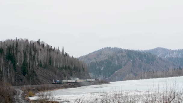 さびた車を持つ古い電車は 春の初めに氷で覆われた川と山々の間を通る鉄道のレールに沿って移動します 古い電車が向かって移動して川岸を通る鉄道 — ストック動画