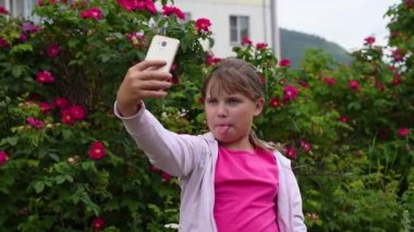 Kız çiçek açan gül bir arka plan bahçede telefonda bir selfie alır. Sarışın saçlı Avrupalı kız akıllı telefonunda kendi fotoğraflarını çekiyor