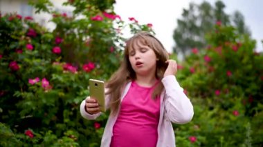 Kız çiçek açan gül bir arka plan bahçede telefonda bir selfie alır. Sarışın saçlı Avrupalı kız akıllı telefonunda kendi fotoğraflarını çekiyor