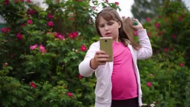 Kız çiçek açan gül bir arka plan bahçede telefonda bir selfie alır. Sarışın saçlı Avrupalı kız akıllı telefonunda kendi fotoğraflarını çekiyor
