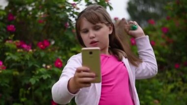 Sarışın saçlı Avrupalı bir kız akıllı telefonunda kendi fotoğraflarını çekiyor. Kız çiçek açan gül bir arka plan bahçede telefonda bir selfie alır