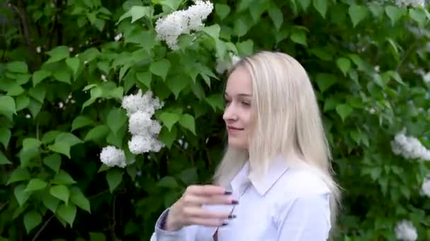 白いライラックの背景に若い女の子は笑顔の髪をまっすぐに 白いライラックの前に長いブロンドの髪を持つ女の子 — ストック動画