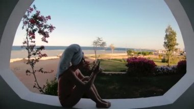 Bir kız pencerenin yanında duruyor ve cep telefonuyla bir telefon numarasını çeviren.