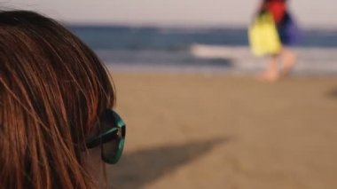 Güneş gözlüklü bir kız denize bakıyor, insanların siluetleri kumsalda yürüyor..