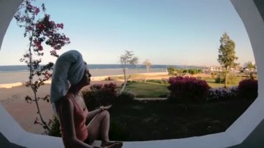 Denize bakan pencere. Bir kız bir kütüğe oturur ve bir cep telefonuna bakar..