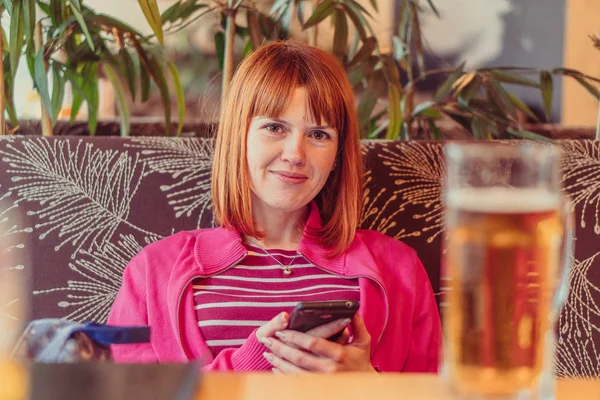 Dívka sedící v hospodě pije pivo a dívá se do telefonu — Stock fotografie