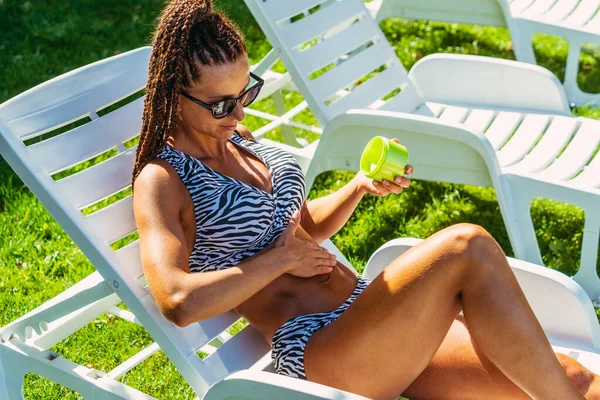 Bronzlaşmış. Mayolu bir kadın güneş kremi sürüyor..