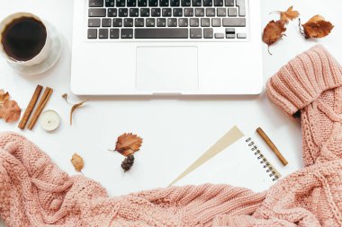 Ofis masası çalışma alanı. Örgü süveter, bir fincan kahve, dizüstü bilgisayar, defter, sonbahar yaprakları, tarçın çubukları, beyaz arka planda mumlar. Sonbahar kompozisyonu. Düz yatış, üst görünüm