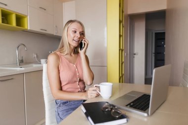 Evde mutfak masasında oturan genç bir kadın cep telefonuyla müşteriyle konuşuyor dizüstü bilgisayara bakıyor şirket müşterilerine danışmanlık yapıyor, iş arkadaşlarıyla sohbet ediyor ortak sorunları çözüyorlar.