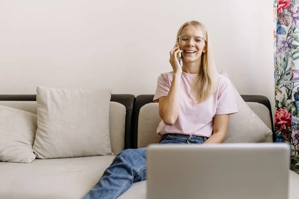 Young woman is sitting relaxed on the sofa talking with client by cell phone call looking at laptop screen provide consultation to corporate customer, chatting with colleague solve common issues.