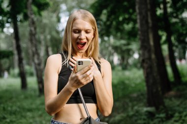 Çekici sarışın kadın elinde akıllı bir telefona bakarken mutlu bir şekilde bağırıyor. İyi haberleri var. Şık kıyafetler giyiyor.