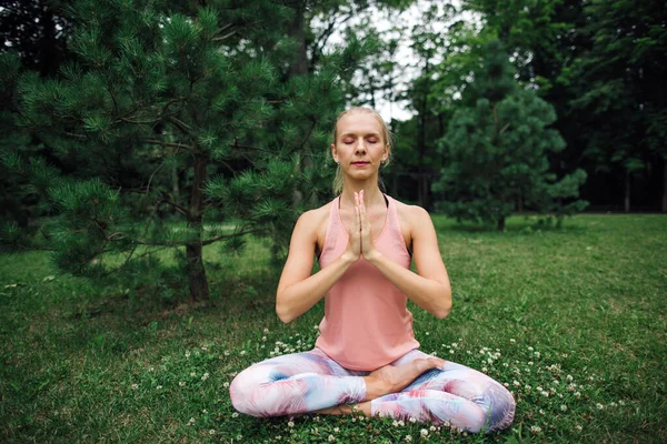 Beautiful young woman seating in Yoga pose of lotus in the park and meditating on green grass