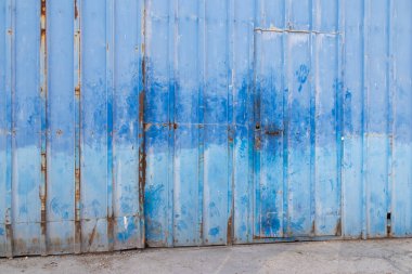 Yıpranmış, el izi, pas ve kabuk bağlayan mavi sürgülü metal kapı desenli, klasik endüstriyel bir zemin oluşturuyor.