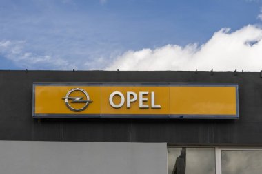 Opel logosu bulutlu bir gökyüzü altında otomotiv endüstrisini ve marka kimliğini temsil eden bir galeri binasında sergileniyor.