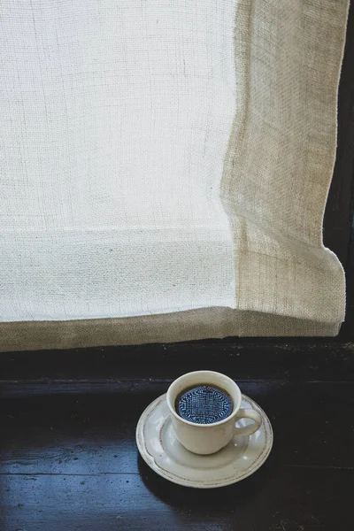 A cup of black coffee with a saucer is on the windowsill with a curtain. Vintage style.