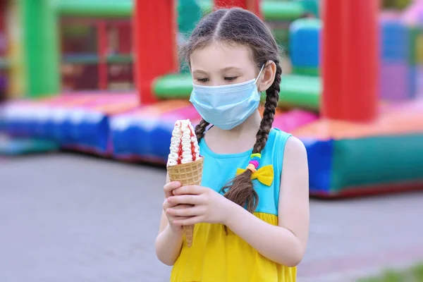 A little girl in a medical mask is sad because she does not allow her to eat ice cream, which she so wants. Coronavirus Protection Concept
