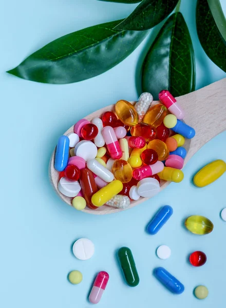 A wooden spoon with multi-colored capsules,health supplements and vitamins with green leaves on a blue background.Supplemental and healthcare product, flat lay