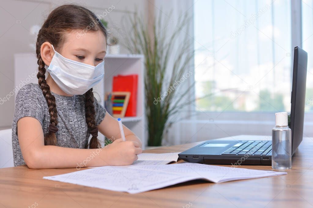 A schoolgirl in a medical mask is doing her homework on a laptop at home or studying in class at a lesson at school.Student safety after covid-19 pandemic. Back to school.Distance learning.