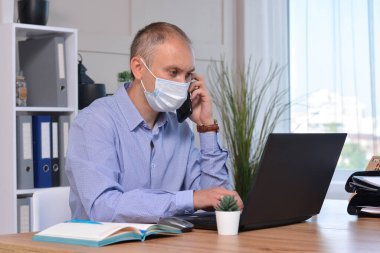 Ofiste tıbbi maske takan bir işadamı kendini korumak için dezenfektan kullanır. Dizüstü bilgisayarda çalışıyor ve telefonda konuşuyor. Coronavirus salgını, Covid 19, mevsimsel grip..