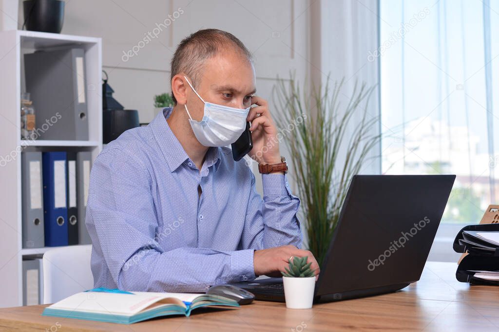 A businessman works in an office wearing a medical mask, uses a disinfectant to protect himself. He works on a laptop and speaks on the phone. Coronavirus pandemic, Covid 19, seasonal flu.