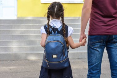 Okula dönelim. Babası küçük kızının elini tutuyor ve okulun arka planında bir sırt çantası var..