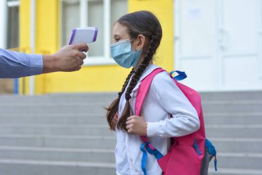 Okulda sıcaklık kontrolü var. Tıp maskesinde çocuk covid-19 salgınında sınıfta. Termometresi anaokulunda ya da okul girişinde olan öğretmen. Sosyal uzaklık. Coronavirus önleme.