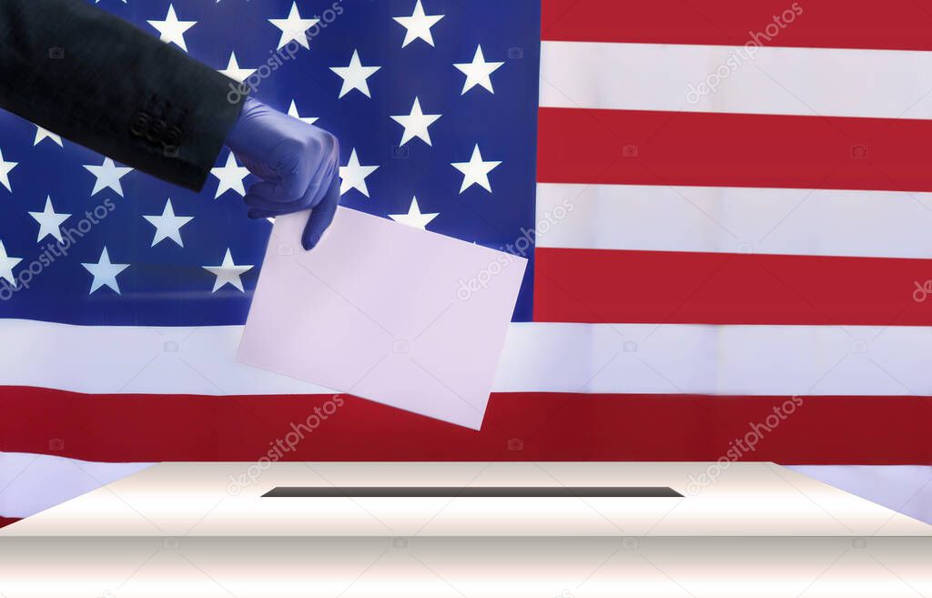 A man in a business suit and blue latex protective gloves puts a ballot paper in a ballot box, against the background of the American flag. US presidential election day during the coronavirus pandemic