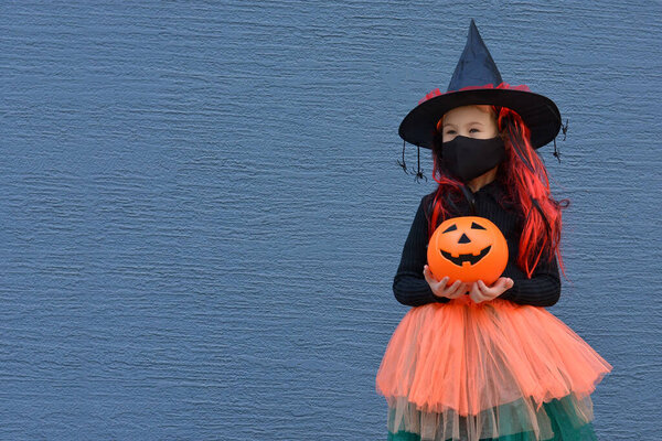 A little girl in a witch costume in a medical mask holding a bucket of pumpkin for sweets on Halloween day against a blue wall. Halloween with safety measures from Covid-19.Copy space for text