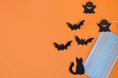 Tıbbi koruyucu maskenin üst görüntüsü, siyah yarasa silueti, hayaletler, turuncu arka plandaki kedi. Yeni bir gerçeklikte 2020 Cadılar Bayramı. Cadılar Bayramı, COVID-19 önleme ve yeni normal kavram. Düz yatıyordu.