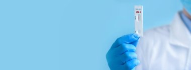 Koruyucu maske ve eldiven takan bir doktor yeni koronavirüsün IgM ve IgG antikorlarını tespit etmek için Covid-19 Yeni Varyant JN1 için hızlı laboratuvar testi gösteriyor. Pozitif sonuç. Pankart