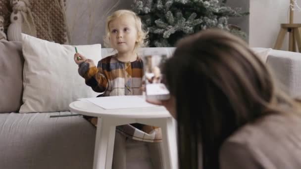 幼い母親は サンタクロースへの手紙を書き 絵を描くスマートフォンで娘の写真やビデオを撮影します クリスマスツリーの背景 — ストック動画